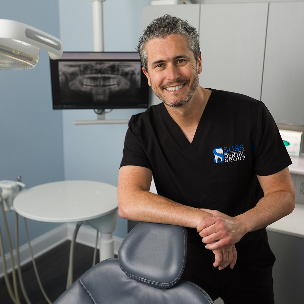 The image shows a man standing in front of dental equipment, smiling slightly, with a backdrop featuring a dental chair and an X-ray machine. He has a stethoscope around his neck and is wearing a black shirt with a logo on it, which appears to be related to dental care or practice.