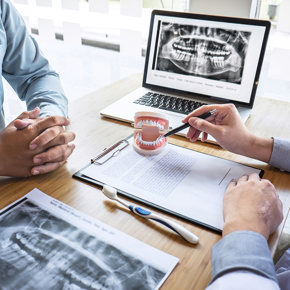 A dental professional is showing an X-ray image to a patient with a laptop, while seated at a table with a clipboard, pen, toothbrush, and other dental equipment.